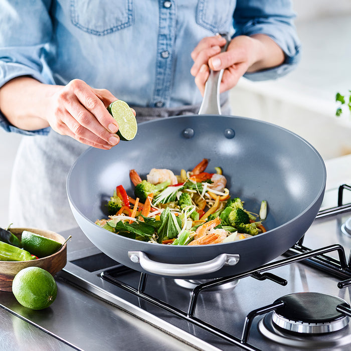 Shrimp and vegetables cooking in a green ceramic nonstick wok, hand squeezing lime over dish in sunlit kitchen.