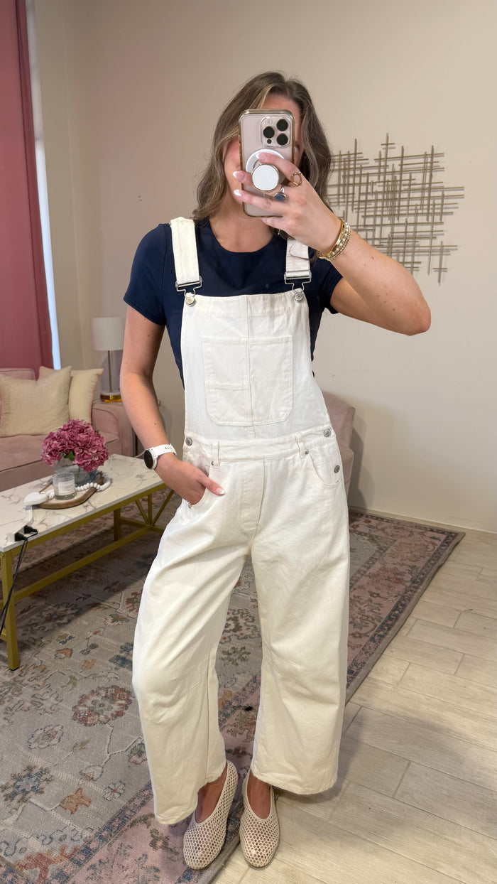 A woman takes a mirror selfie wearing cream wide-leg denim overalls over a navy blue t-shirt, paired with beige slip-on shoes. The setting is a cozy living room with a patterned rug, sofa, and decorative wall art.