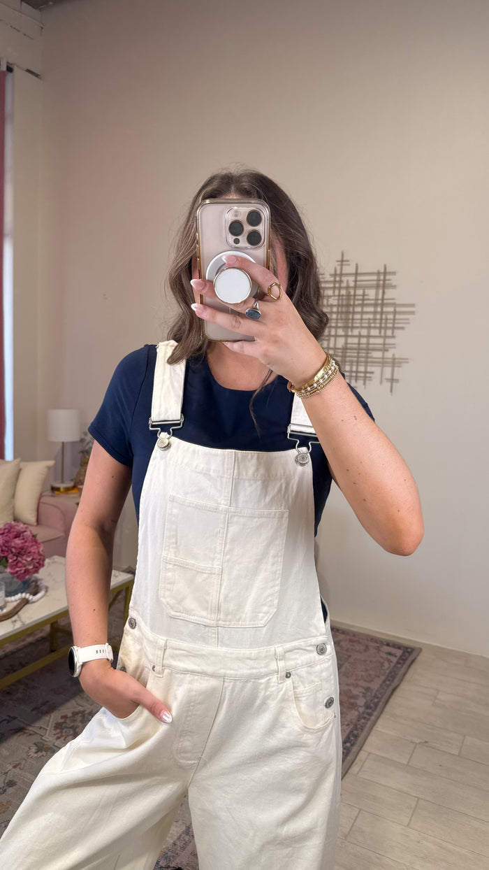 A woman takes a mirror selfie wearing cream-colored denim overalls over a navy blue t-shirt in a cozy living room setting.