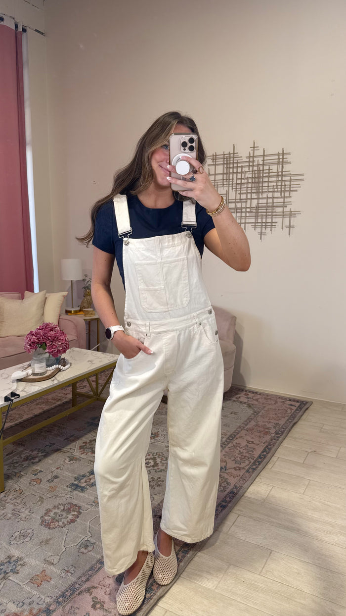 A woman takes a mirror selfie in a living room, wearing a navy blue t-shirt and cream-colored wide-leg denim overalls. She is standing on a patterned area rug with a sofa and decorative wall art in the background.