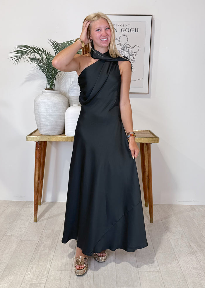 A woman modeling a black halter-neck maxi dress with a draped, asymmetrical design, standing in a brightly lit room with a wooden console table and framed art in the background.