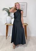 A woman modeling a black halter-neck maxi dress with a draped, asymmetrical design, standing in a brightly lit room with a wooden console table and framed art in the background.