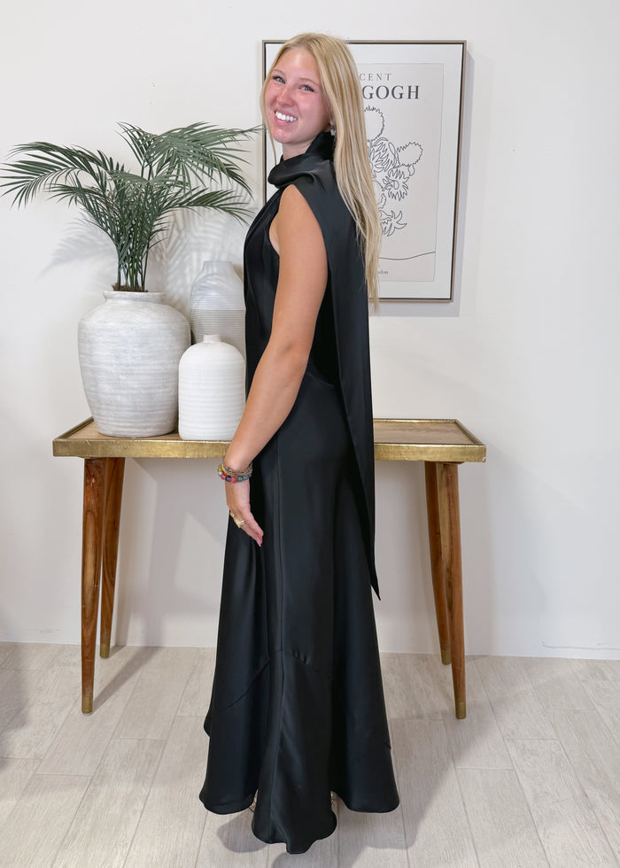 A woman modeling a sleeveless black maxi dress with a high draped collar and a long flowing cape-like back drape, standing in a bright room with a console table, white vases, and a framed print.