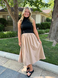 A woman stands outdoors on a sidewalk in front of a house and trees, wearing a sleeveless black top and a flowing beige midi skirt with black sandals.
