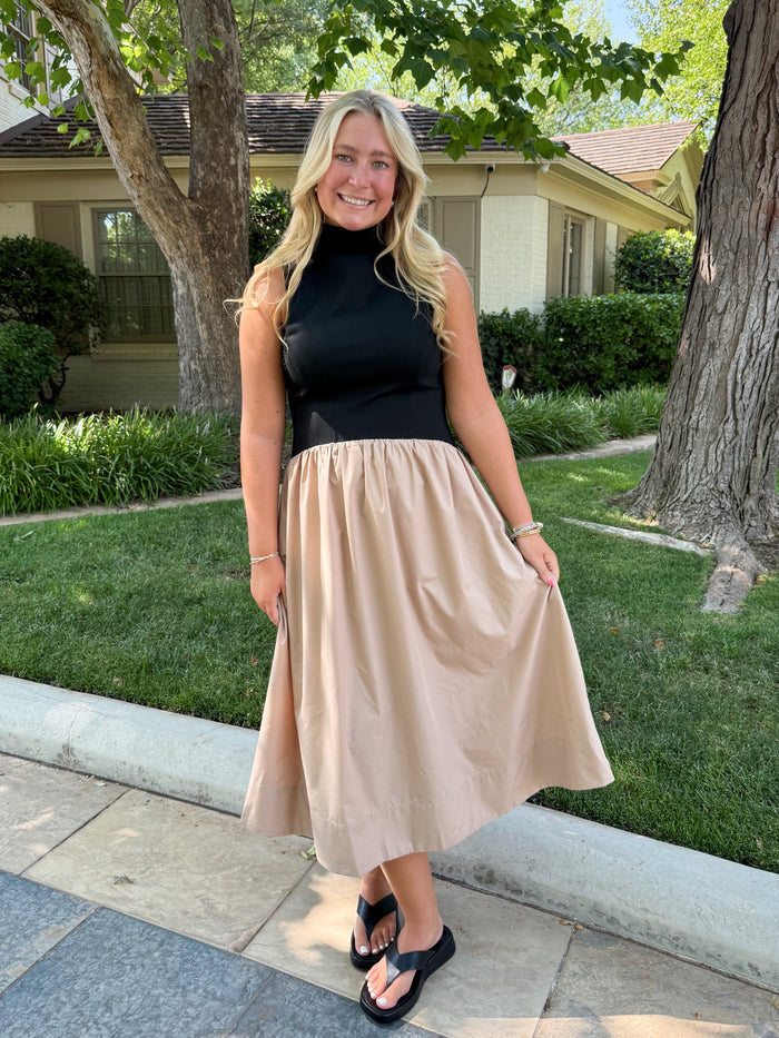 A woman stands outdoors on a sidewalk in front of a house and trees, wearing a sleeveless black top and a flowing beige midi skirt with black sandals.