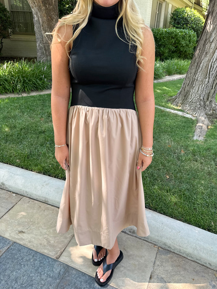 A woman wearing a sleeveless black and beige two-tone midi dress with a high neck and gathered waist, standing outdoors on a stone walkway with grass and trees in the background.