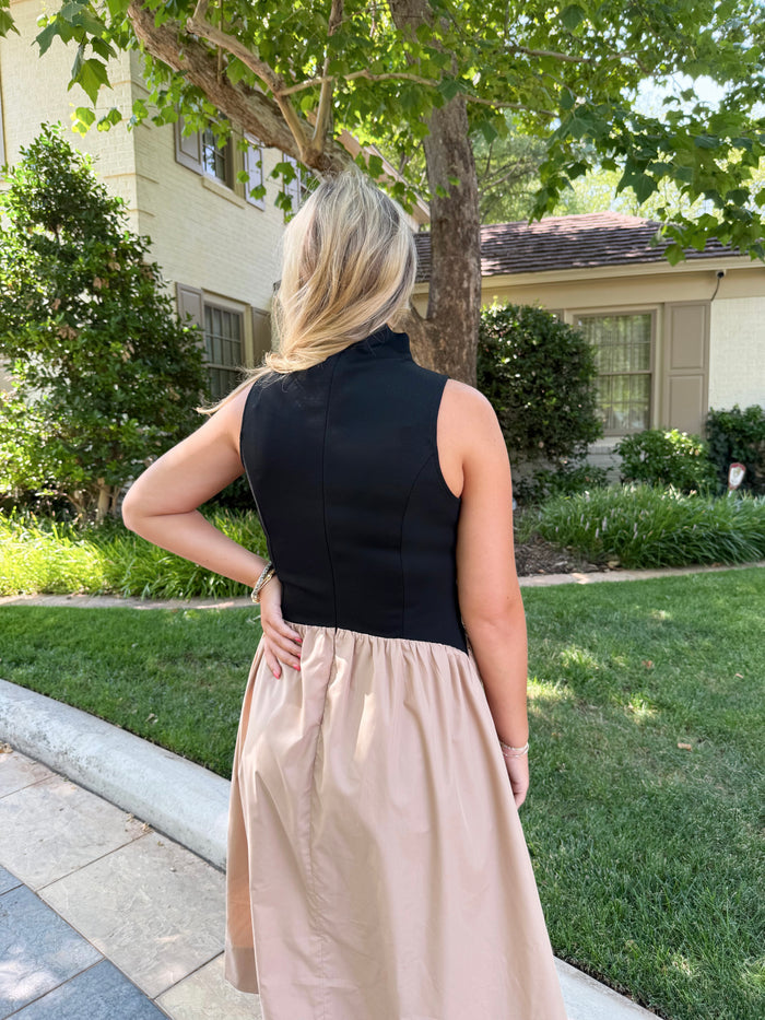 A woman viewed from behind wearing a sleeveless high-neck dress with a black bodice and a flowing blush-beige skirt, standing outdoors on a sidewalk with grass, trees, and a house in the background.