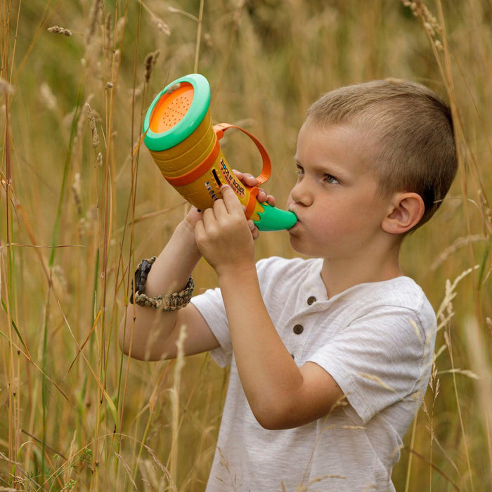 A young boy in a white shirt stands among tall grasses, blowing into the Outdoor Discovery Hoot N Holler Animal Caller, enjoying creative outdoor play.