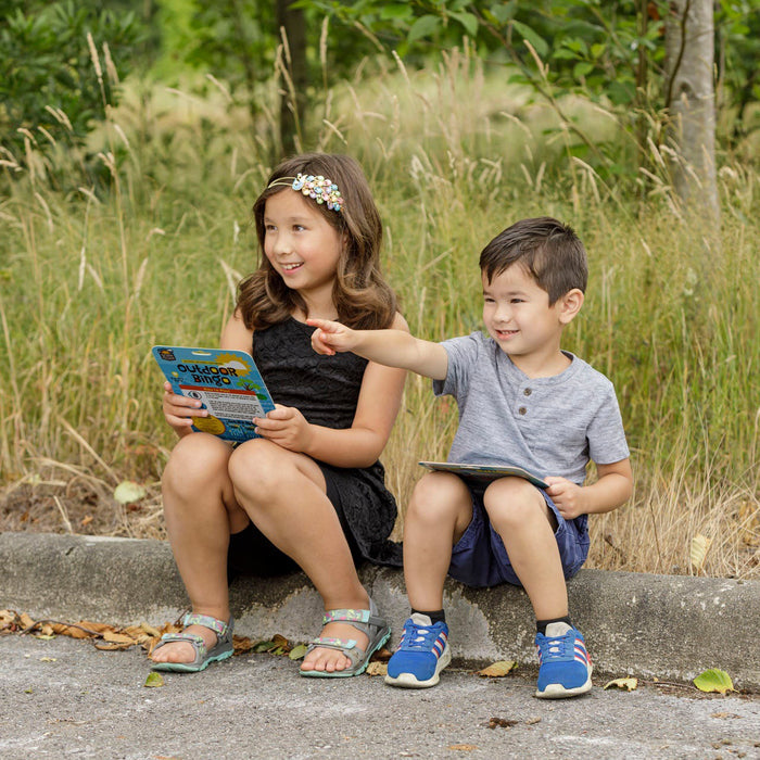 Two young children sit on a curb outdoors; the girl reads while the boy points off camera. Their Outdoor Discovery Outdoor Bingo 4 Pack rests beside them, surrounded by tall grass and trees.