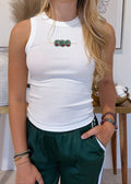 A young woman with blonde hair wearing a white ribbed tank top with a small green and red graphic on the chest, paired with dark green athletic pants, standing in a styled indoor setting.