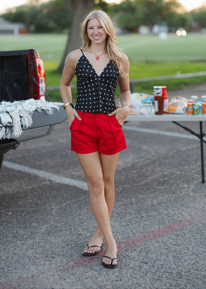 A woman stands outdoors in a parking lot next to a truck and a folding table, wearing a black sleeveless top with white polka dots and bright red shorts.