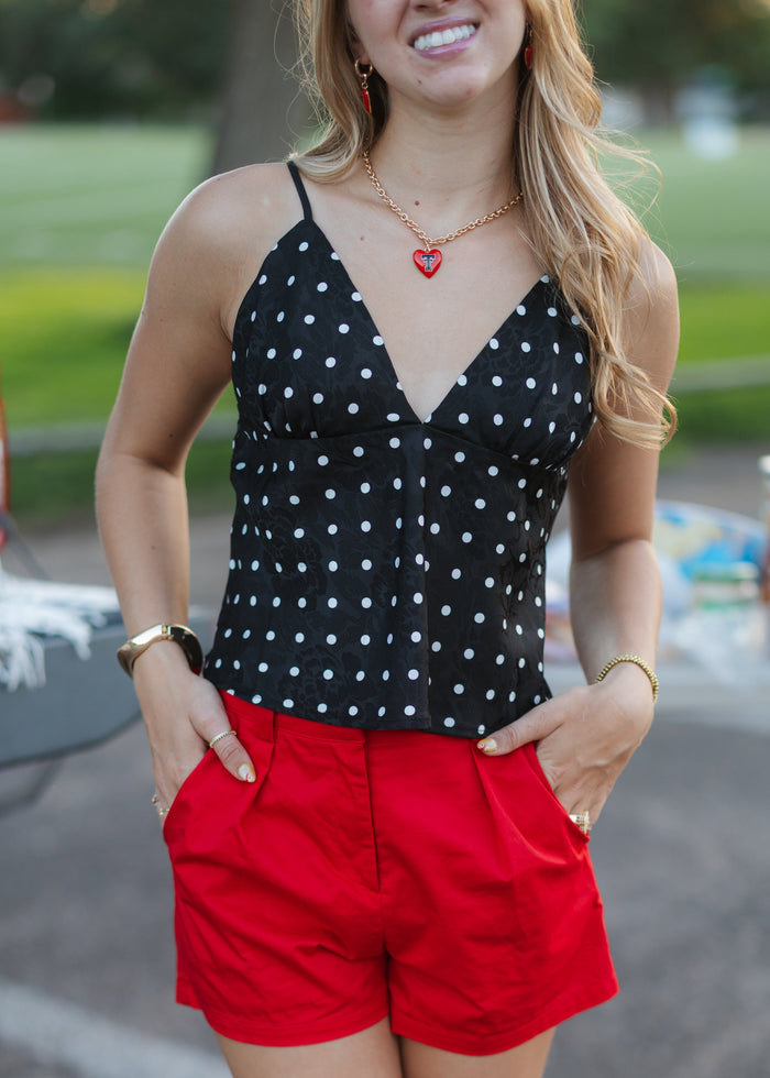 A woman standing outdoors on a paved surface wearing a black camisole top with white polka dots and bright red shorts. She is smiling and posing with her hands in her pockets, with a picnic setup and a vehicle visible in the background.