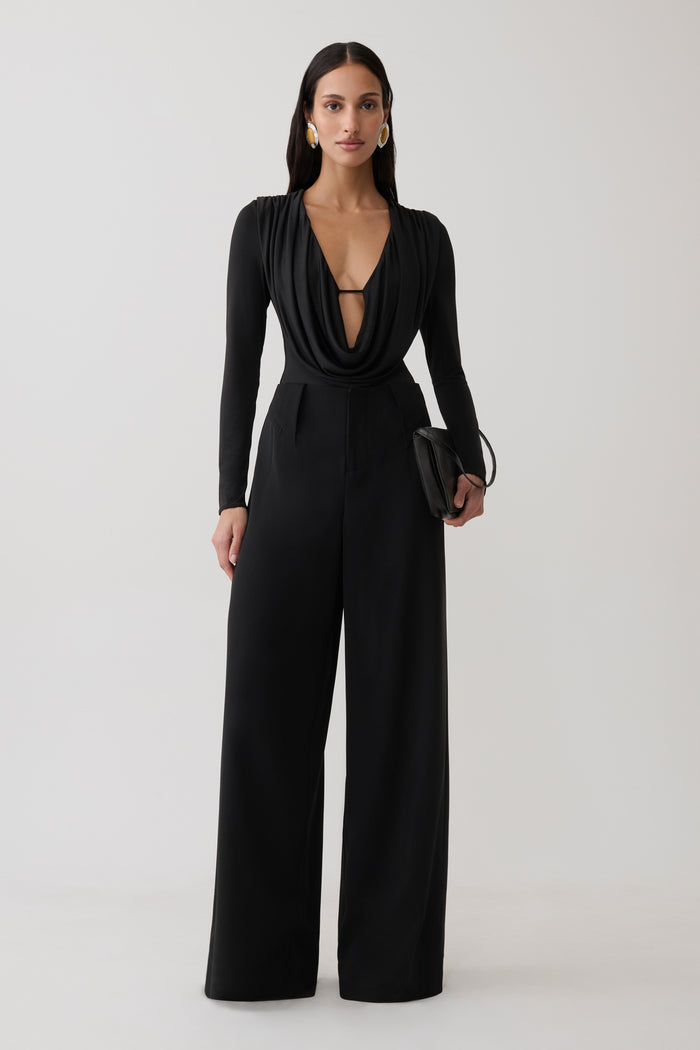 Woman wearing black cowl long sleeve slinky bodysuit, with black pants.
