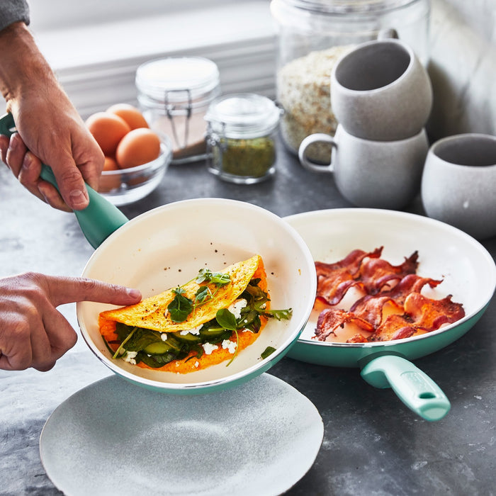 Turquoise ceramic pan from a 16-piece cookware set holding a folded omelette with greens, bacon nearby.
