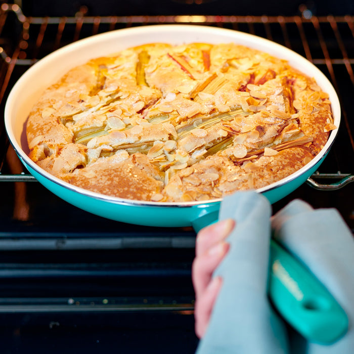 Turquoise ceramic nonstick pan with almond rhubarb cake held in front of an open oven.