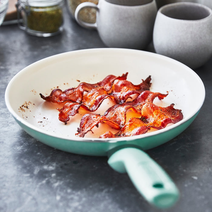 Bacon strips cooking in a turquoise-handled ceramic nonstick pan from a 16-piece set on a gray countertop.