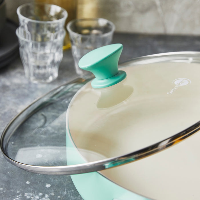 Turquoise ceramic nonstick 16-piece cookware set with glass lid on a gray countertop next to clear glasses.