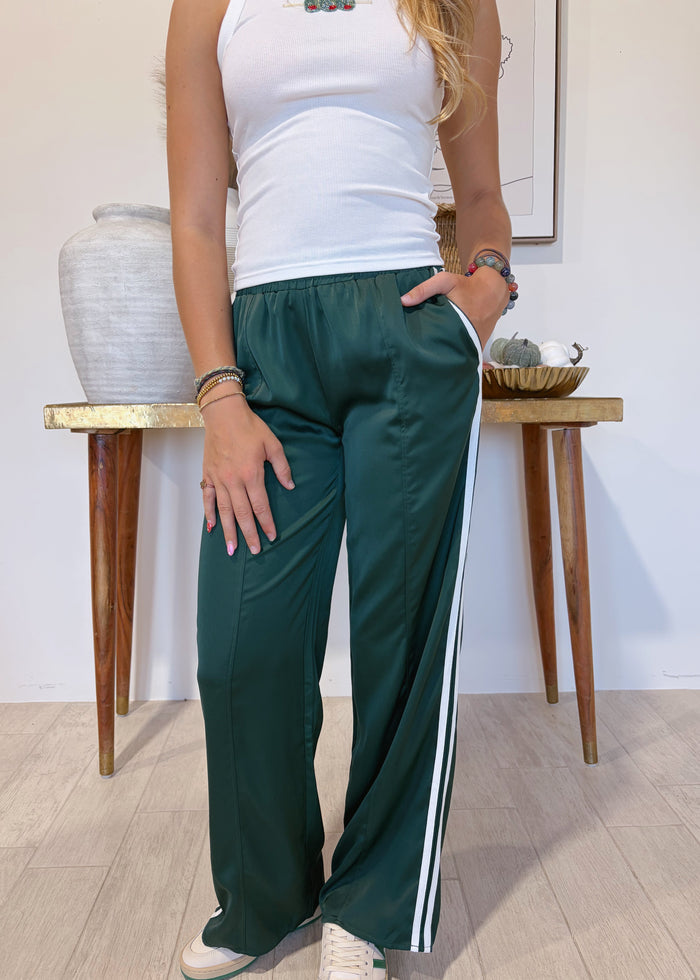 A person wearing dark green wide-leg track pants with white side stripes, paired with a white tank top and white sneakers, standing in a room with a wooden table and vase in the background.
