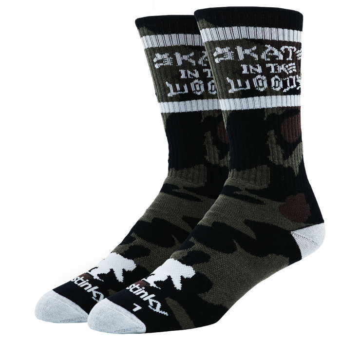 A pair of crew socks featuring a black and olive green camouflage pattern, with white text reading 'SKATE IN THE WOODS' on the upper cuff and white accents on the toes and heels, set against a plain white background.