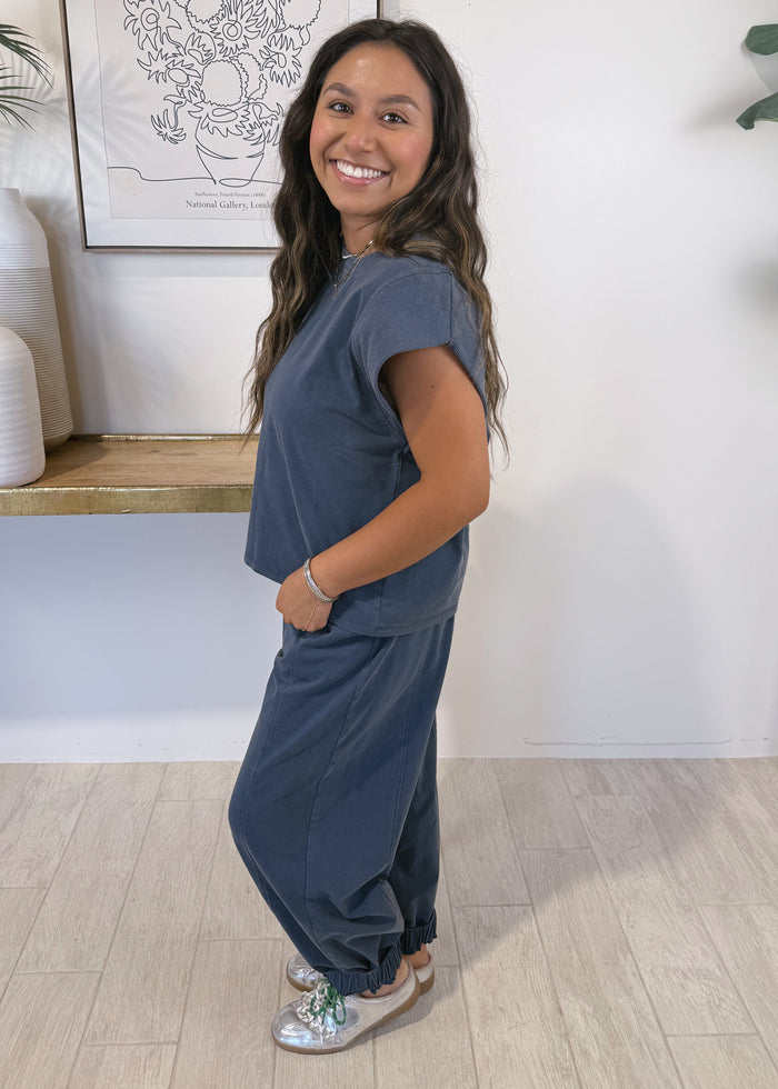 A woman modeling a matching slate blue lounge set consisting of a short-sleeve top and matching pants, standing in a brightly lit room with a white wall, wooden floor, and decorative plants.