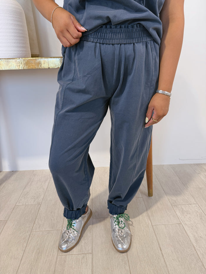 A person wearing loose, faded blue-gray jogger-style pants with an elastic waistband and gathered cuffs, paired with a matching top and metallic silver shoes, standing indoors on a light wood floor.