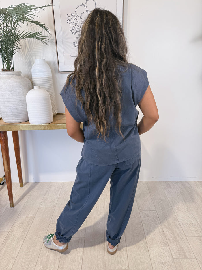 A woman viewed from behind wearing a matching slate blue scrub top and scrub pants set, standing in a bright room with light wood floors and white ceramic vases.