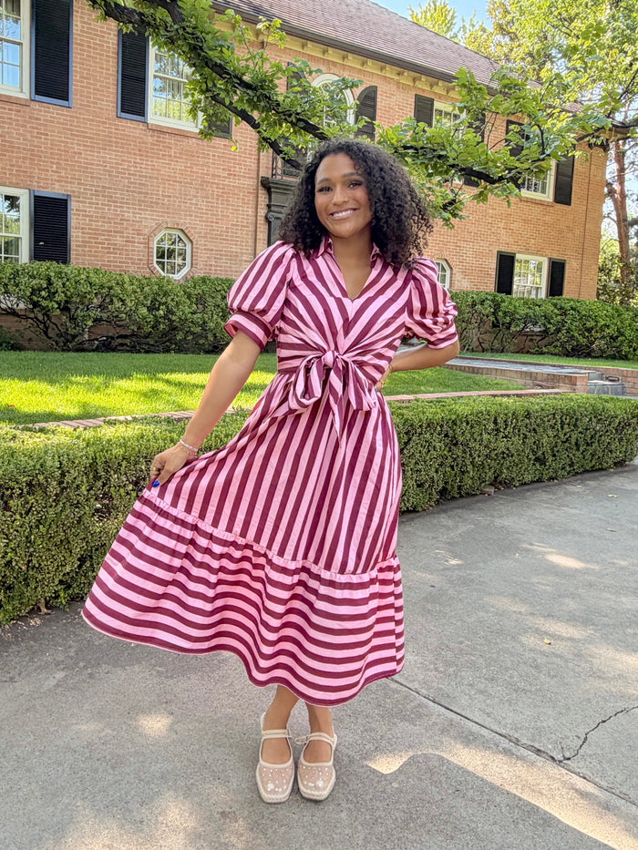 A woman wearing a pink and white vertically striped midi dress with puff sleeves and a tie waist, standing outdoors on a paved walkway in front of a brick building and green hedges.
