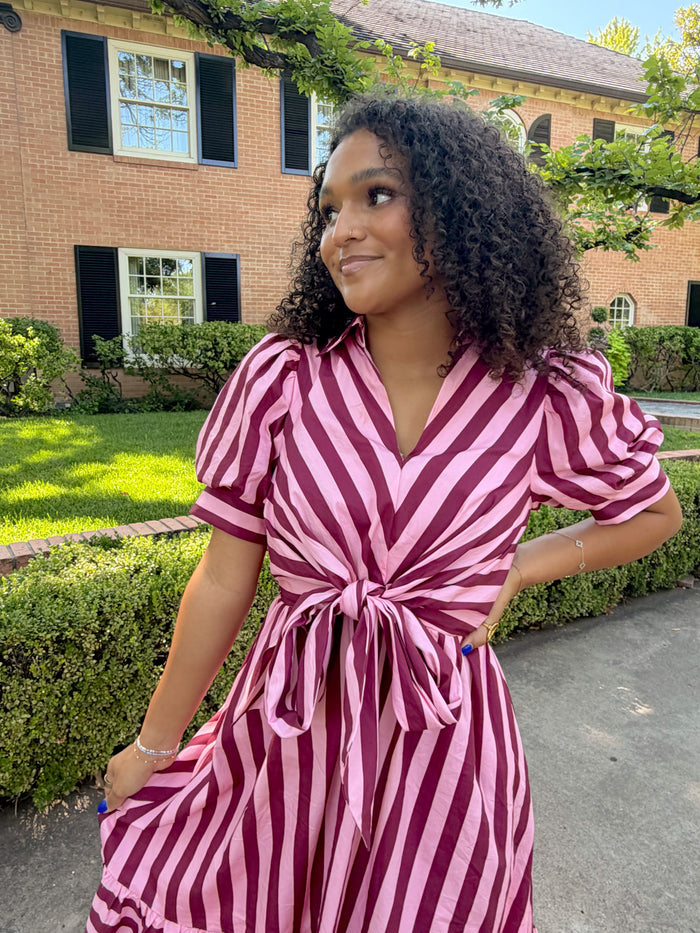 A woman wearing a pink and burgundy striped dress with puff sleeves and a tie waist, standing outdoors in front of a brick house.