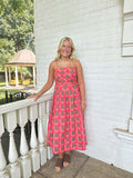 A woman stands on a porch wearing a bright pink sleeveless midi dress with a geometric lattice pattern and cutout details. The background features a white brick wall, a white railing, and a small gazebo in the distance.