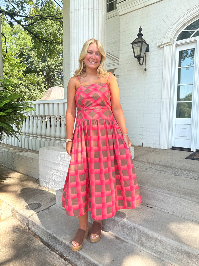 A woman stands on the concrete steps of a white house wearing a sleeveless pink and brown grid-patterned midi dress with sheer mesh panels.