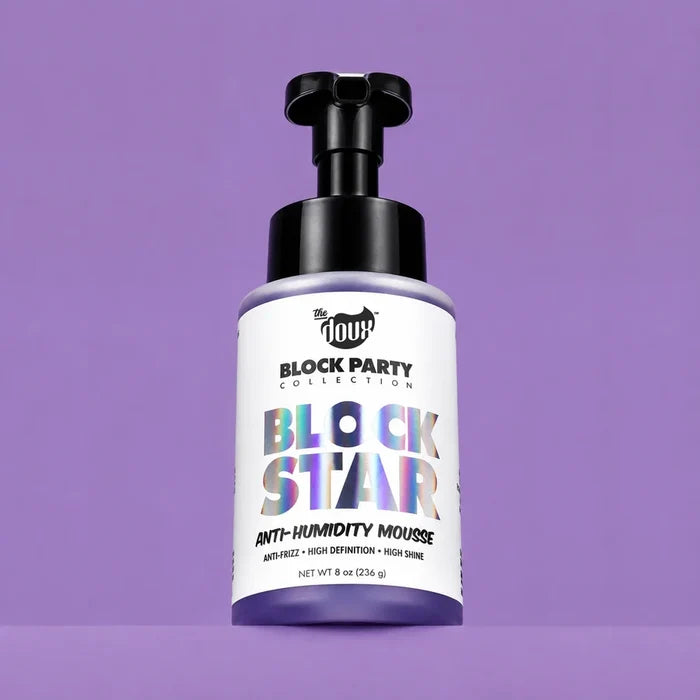 A bottle of The Doux Block Party Collection Block Star Anti-Humidity Mousse, featuring a white label with holographic lettering and a black pump dispenser, set against a solid purple background.