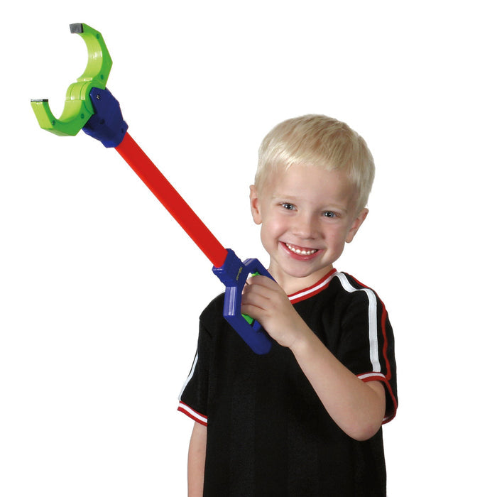 A smiling young blond boy holds up a Toysmith Galaxy Grabber, a colorful toy robot claw by Toysmith with a red handle and green claw, standing against a plain white background.