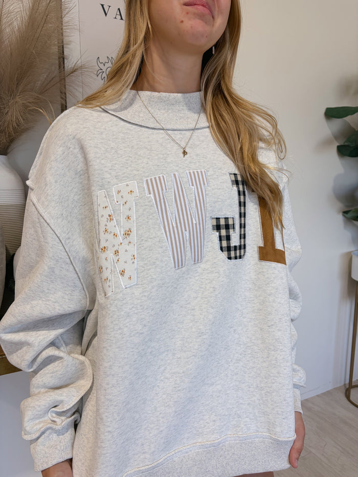 A woman wearing a light heather gray sweatshirt with a large patchwork letter 'W' graphic on the front, styled with a gold necklace in an indoor setting.