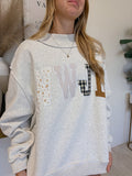 A woman wearing a light heather gray sweatshirt with a large patchwork letter 'W' graphic on the front, styled with a gold necklace in an indoor setting.