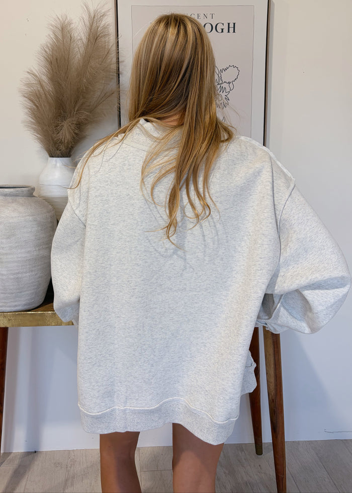 A woman with long blonde hair is shown from behind wearing an oversized light gray heather sweatshirt in a styled indoor setting with a wooden table, vases, and framed wall art.