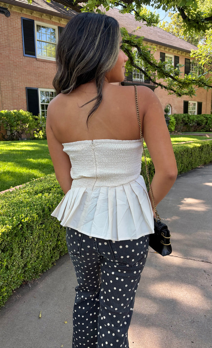 A woman photographed from behind outdoors, wearing a white strapless peplum-style top and black-and-white polka dot pants, standing on a sidewalk in front of a brick building and green hedges.