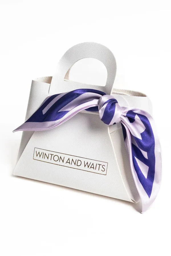 A white paper gift bag with a handle, featuring a purple and white patterned silk scarf tied around the middle and a 'WINTON AND WAITS' logo printed on the front, set against a plain white background.