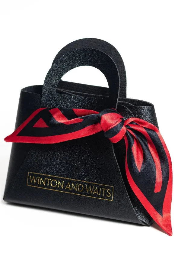 A black textured leather handbag with a top handle, featuring a red and black silk scarf tied around the waist and gold foil branding that reads 'WINTON AND WAITS'.